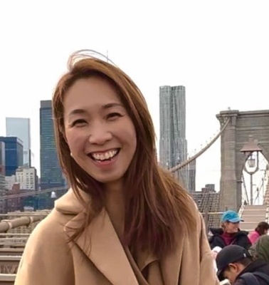 A smiling woman in a beige coat standing in front of the Brooklyn Bridge with the New York City skyline in the background.