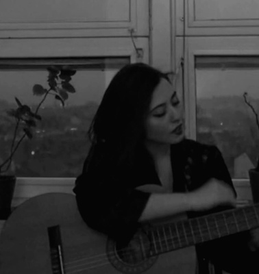 A woman playing an acoustic guitar by a window, with potted plants nearby in a dimly lit room.