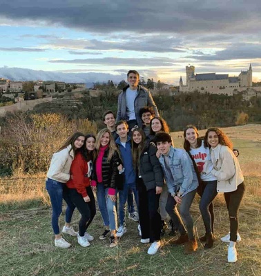 A group of young people posing for a photo in front of a scenic landscape during sunset, with historical buildings in the background.
