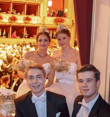 Four young adults in formal attire at a sophisticated event, posing with flowers in a theater-like setting with an audience in the background.