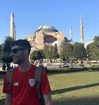 A man in a red t-shirt stands in front of the Hagia Sophia with a clear blue sky above.