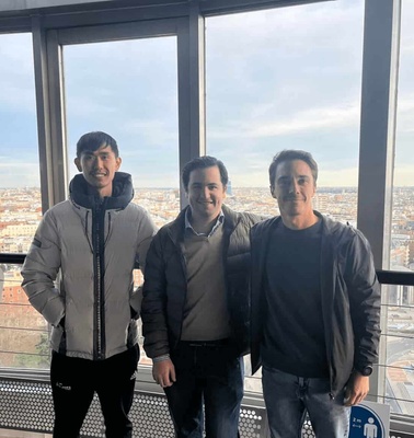 Three men smiling inside a glass-walled room with a cityscape in the background.