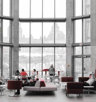 A modern lobby with large windows, red furniture, and people sitting and talking with a city view in the background.
