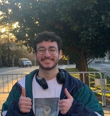 A young man with glasses and headphones around his neck is smiling and giving a thumbs up in front of a tree-lined street and buildings.