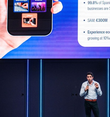 A presenter discusses business insights at IE University with a large screen displaying data and a phone showing images.
