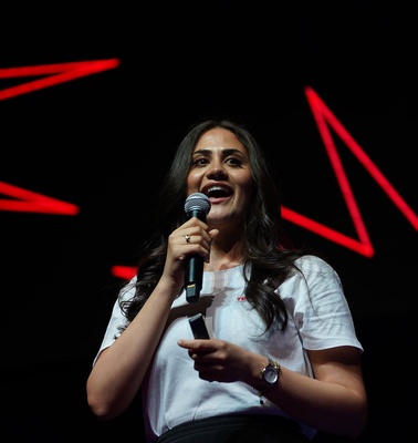 A woman is speaking confidently into a microphone on stage with red star-like decorations in the background.