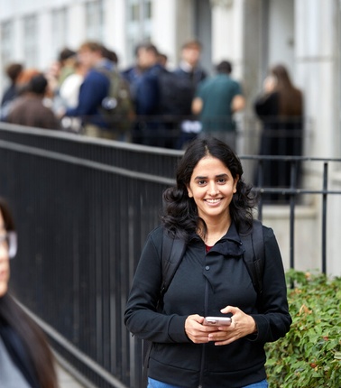 A smiling woman with a phone stands in the foreground with a blurred queue of people in the background.