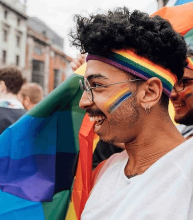 A joyful young man with curly hair wearing a rainbow headband and face paint at a Pride parade, smiling while holding a rainbow flag.