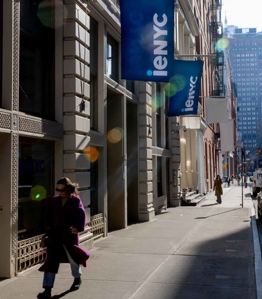 A woman walks along a sunny city street lined with buildings and banners.