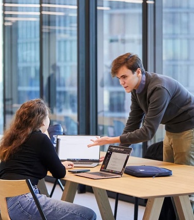 A man is explaining something to a woman at a modern workspace with laptops.