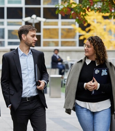 Two professionals are walking and engaging in conversation outdoors in an urban setting.