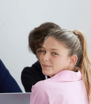 A group of young adults in a casual business meeting or classroom setting, one woman looking back at the camera.