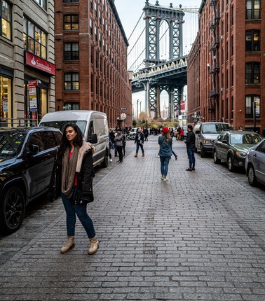 A woman stands on a cobblestone street lined with red brick buildings, with the Manhattan Bridge visible in the background.