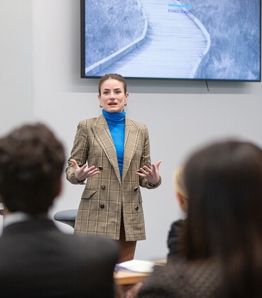 A woman in a checkered blazer is delivering a presentation to an audience.