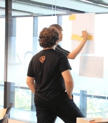 Two individuals are engaged in a collaborative work session, with one writing on a board while the other observes.