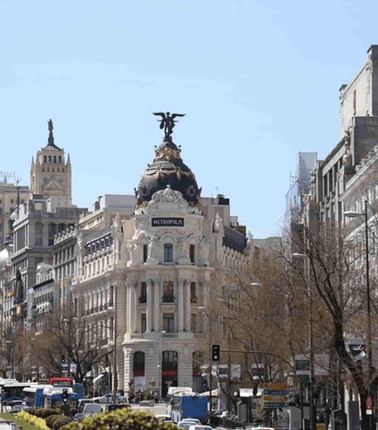 A busy urban street scene featuring classic architecture and a statue on top of a building, under a clear blue sky.