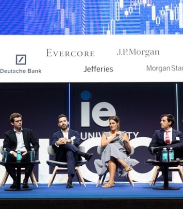 A panel of six professionals, comprising both men and women, are seated on stage at a business conference, with logos of various corporations in the background.