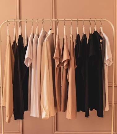 A collection of neatly organized clothes in shades of black, white, and beige on a metal rack against a soft pink background.