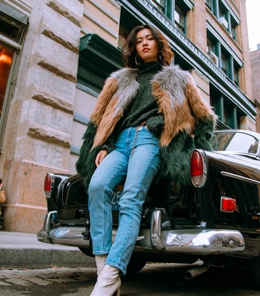 A woman in a chic fur coat and jeans sits on the back of a vintage car on a city street.