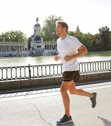 A man in a white t-shirt and black shorts jogging in a park with a monument and fountain in the background.