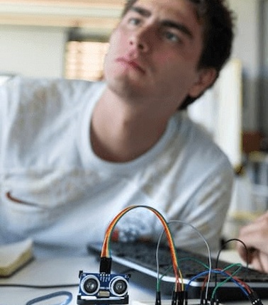 A young man is focused, looking off into the distance while a robotic prototype with wires is placed on a table in front of him.