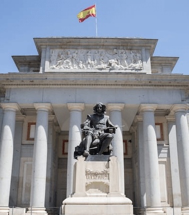 A statue of a notable figure stands in front of a grand building featuring classical architecture and a Spanish flag.