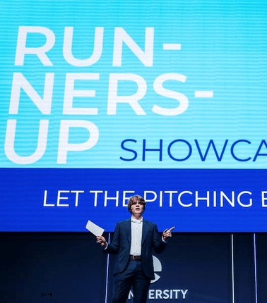 A presenter on stage is introducing a pitching event at a showcase.
