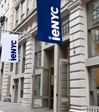 A city street with modern buildings displaying various flags, including one marked 'IENYC'.