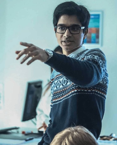 A man in a patterned sweater is gesturing while teaching in a classroom.