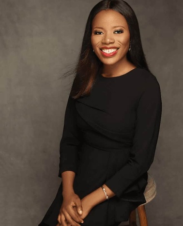 A smiling woman in a black dress sits on a stool against a grey backdrop.