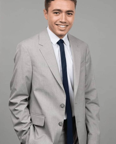 A smiling man in a gray suit with a white shirt and blue tie standing against a gray background.