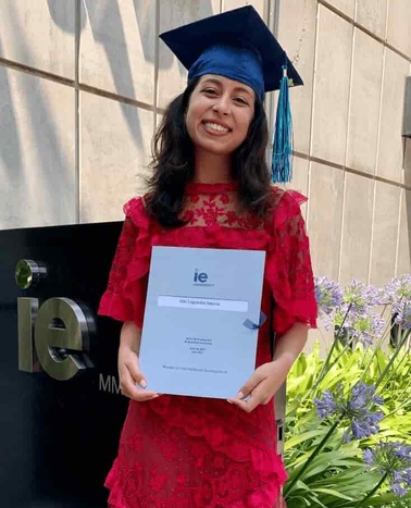 A young woman in a red dress and graduation cap, holding a diploma, smiling at an outdoor graduation ceremony.