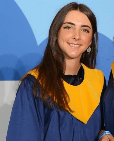 A young woman in a blue and yellow graduation gown smiling at the camera.