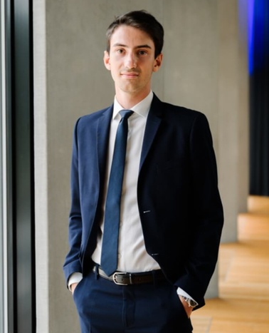 A young man in a business suit standing confidently in a modern office environment.