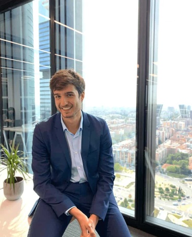 A young man in a blue suit smiling, seated next to a large window overlooking a city.