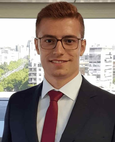 A young man in a business suit with a red tie stands smiling in an office.