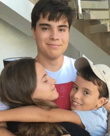 Three young people, likely siblings, smiling and hugging each other indoors.