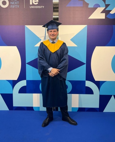 A graduate in a blue and yellow robe stands smiling in front of a colorful 'IE University' backdrop celebrating 'Next Fifty Years'.