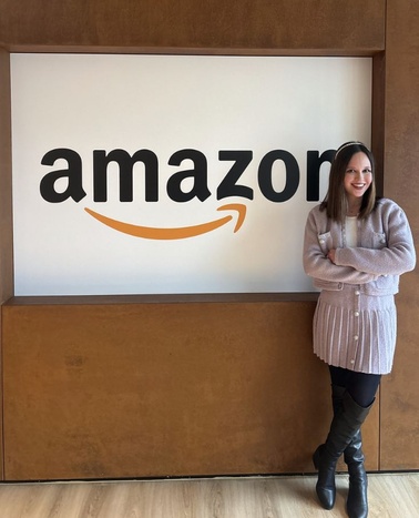 A woman is standing next to the Amazon logo, smiling and dressed in a light sweater and boots.