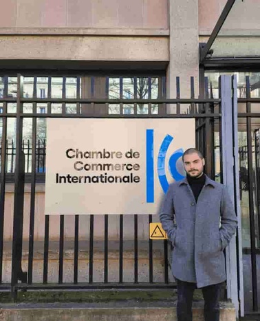 A man in a gray coat standing in front of the Chambre de Commerce Internationale sign mounted on a fence.
