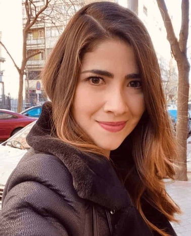 A portrait of a smiling woman with long brown hair wearing a dark jacket, outdoors.