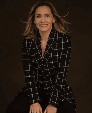 A woman in a plaid blazer smiles while sitting against a dark background.