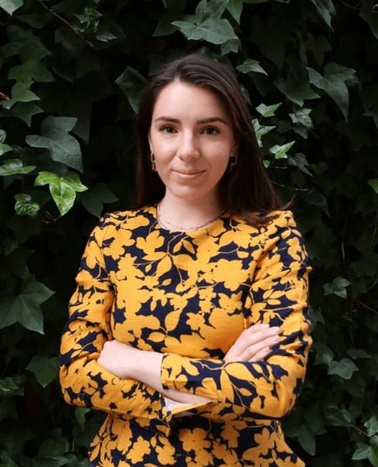 A woman in a yellow and black floral dress stands with her arms crossed in front of a leafy green background.