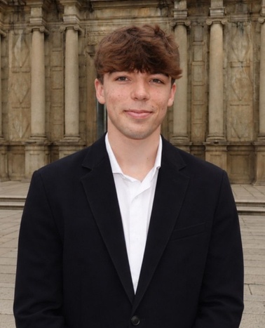 A young man in a black suit jacket and white shirt stands in front of a historic building.