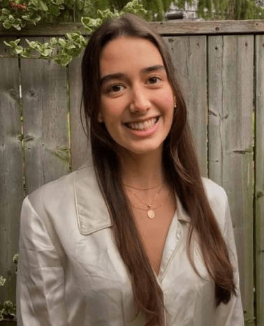 A young woman smiling in a garden, wearing a beige blouse.