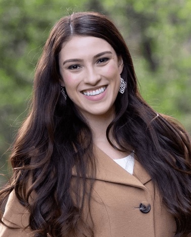 A smiling woman with long brown hair wearing a beige coat and diamond earrings outdoors.