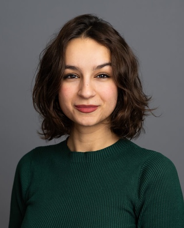 Portrait of a smiling young woman with short curly hair wearing a green sweater against a gray background.