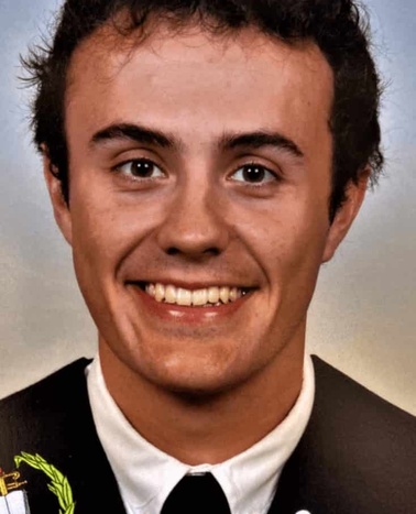 A portrait of a young man smiling, wearing a black suit with a badge and embroidered patch on the jacket.