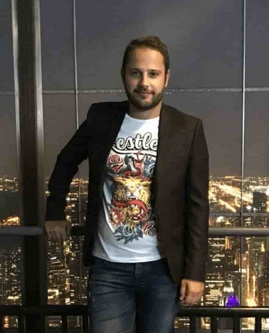 A man in a blazer and printed t-shirt stands in front of a cityscape at night.