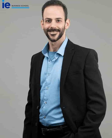 A man in a business suit with a light blue shirt and black blazer stands smiling at the camera.